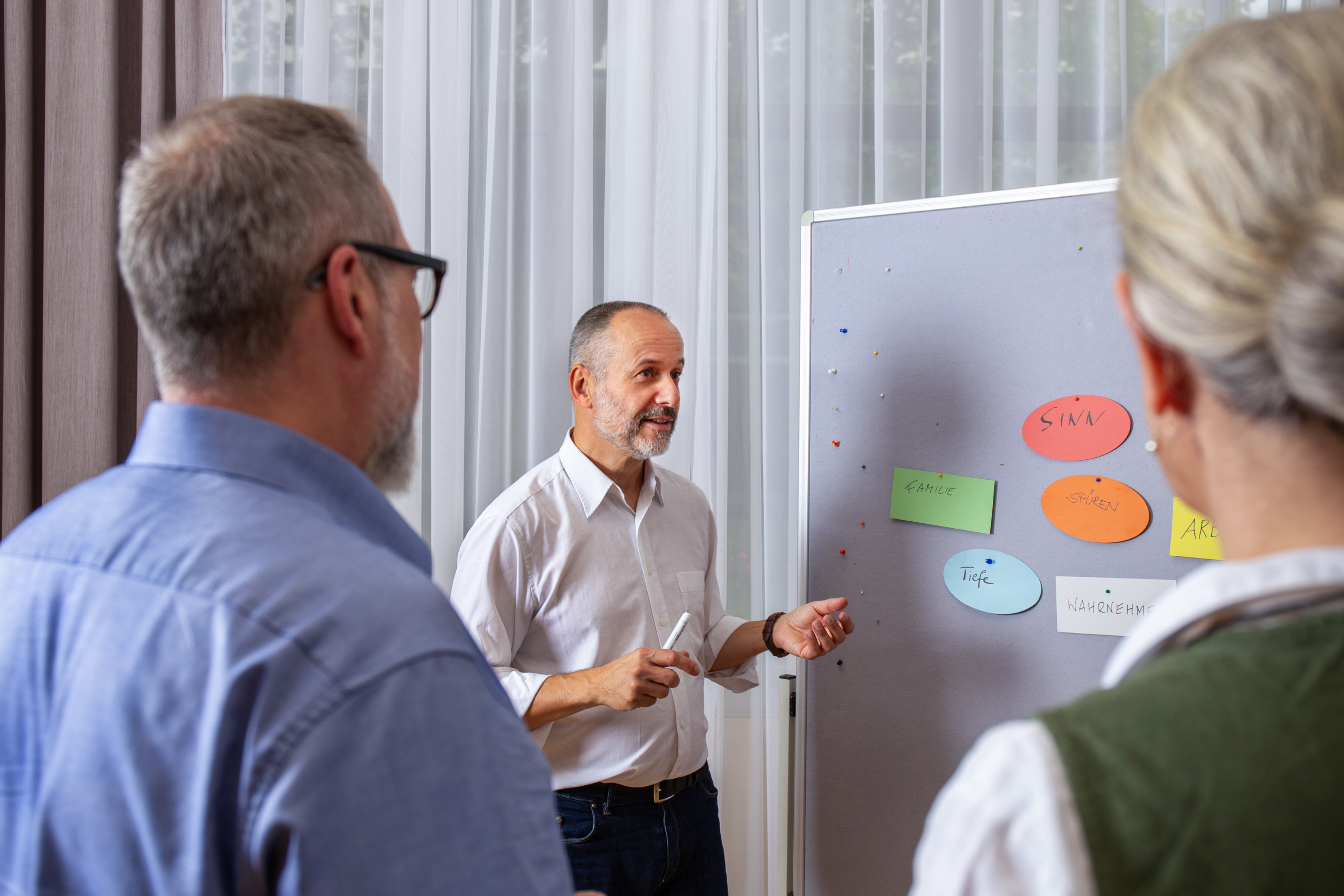 Eine Person erkl&auml;rt zwei Personen etwas. Auf der Pinwand sind bunte K&auml;rtchen. 